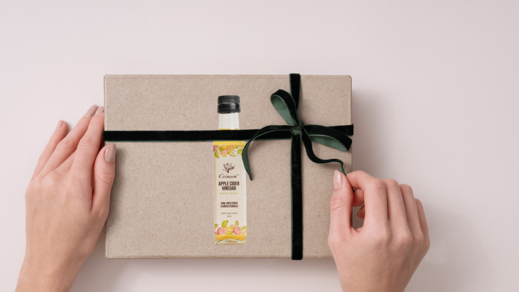 A gift box wrapped in brown paper and tied with a green velvet ribbon, featuring a single bottle of Crimson Apple Cider Vinegar on its cover.