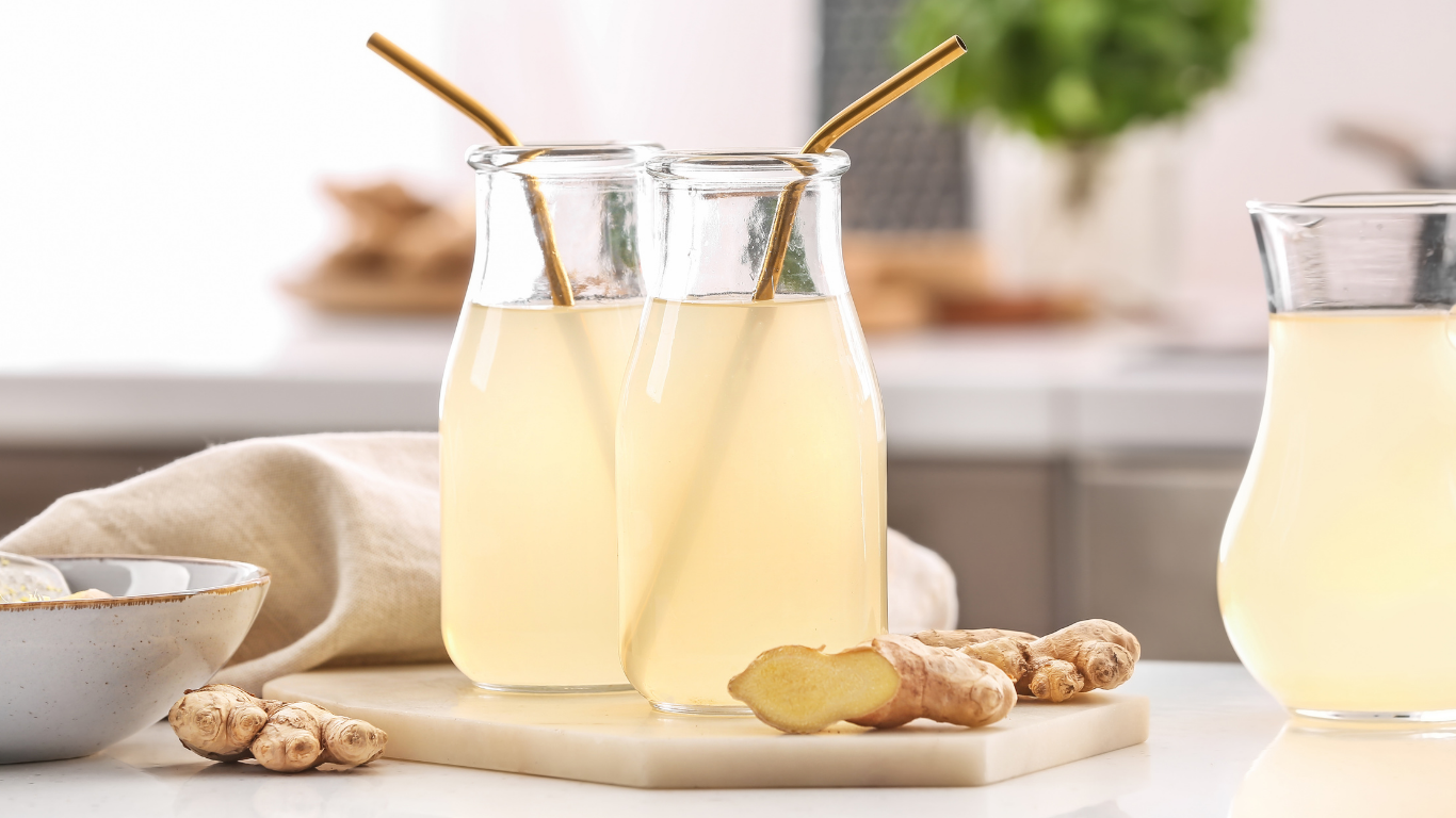 Two small bottles of a light yellow ginger drink with golden straws, alongside fresh ginger roots on a marble board in a bright kitchen setting.