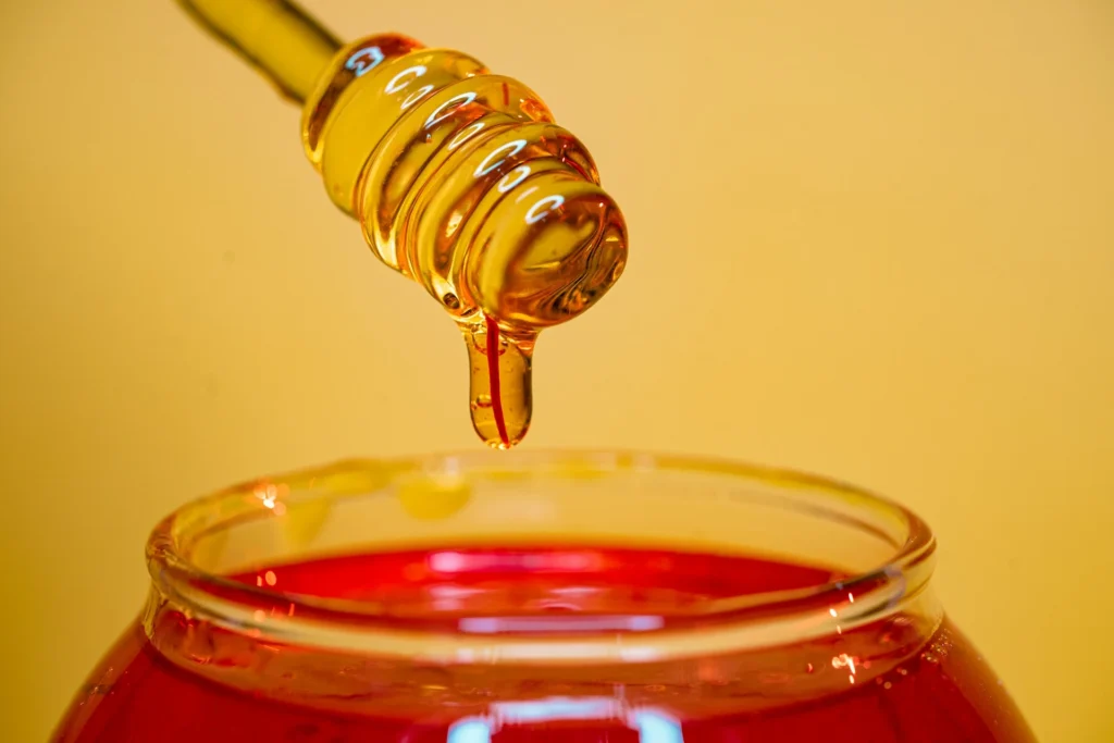 Golden honey dripping from a wooden honey dipper into a glass jar.
