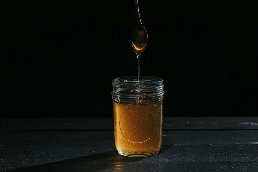 Honey dripping from a spoon into a glass jar against a dark background.